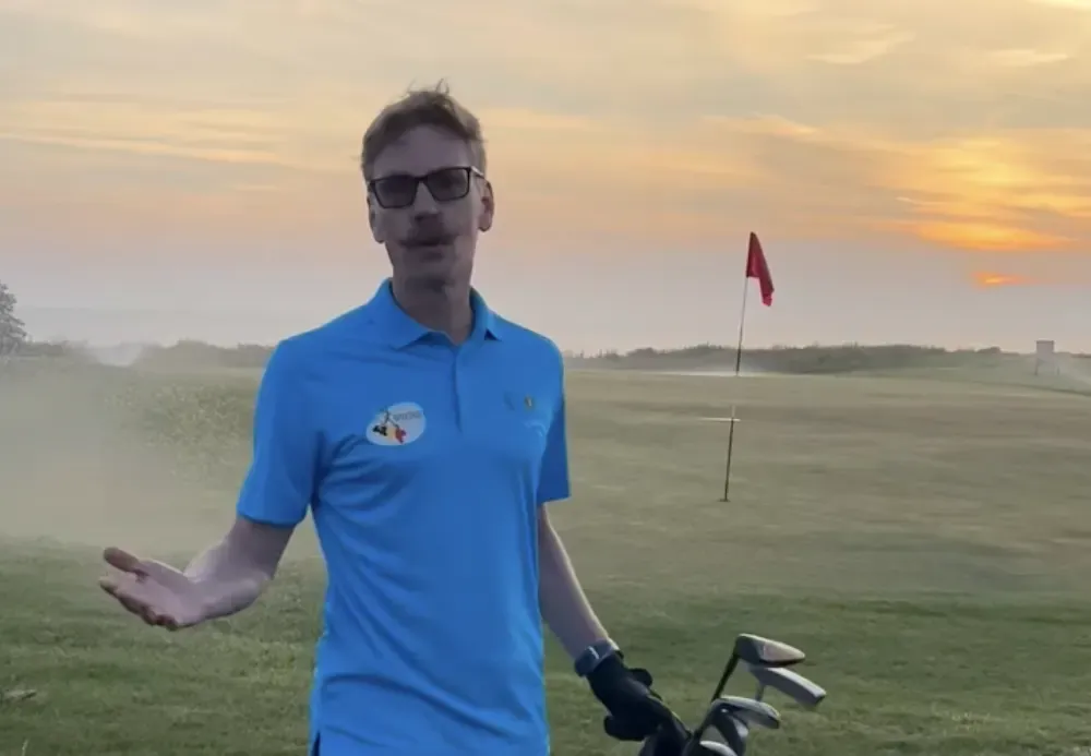 A Scandinavian Open competitor carrying golf clubs beside a green at sunset.