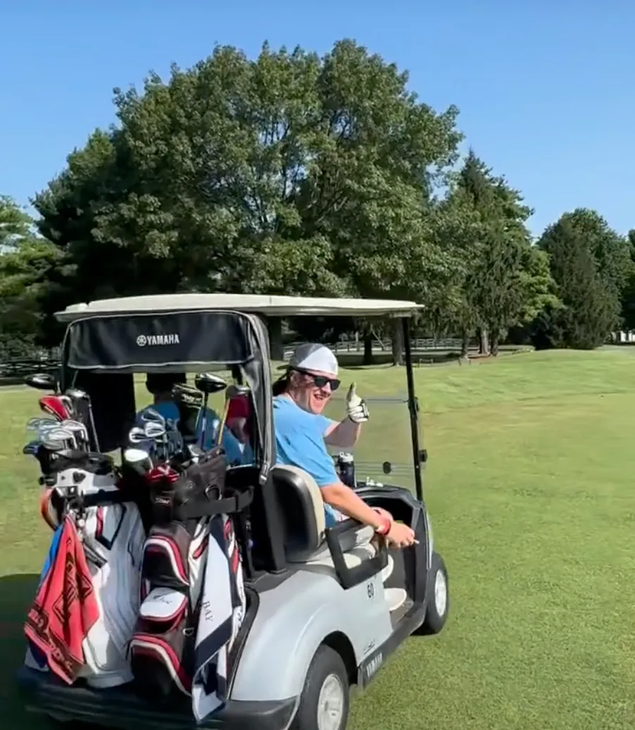 A golfer gives a thumbs-up from a golf cart loaded with clubs on the course.