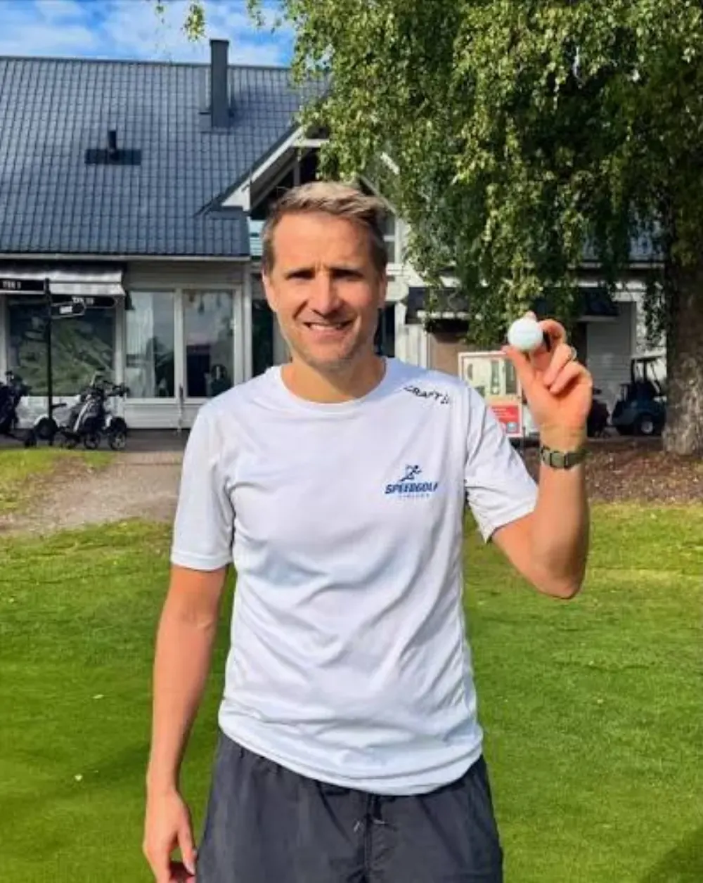 A speedgolfer in a white shirt holds up a golf ball outside a clubhouse.